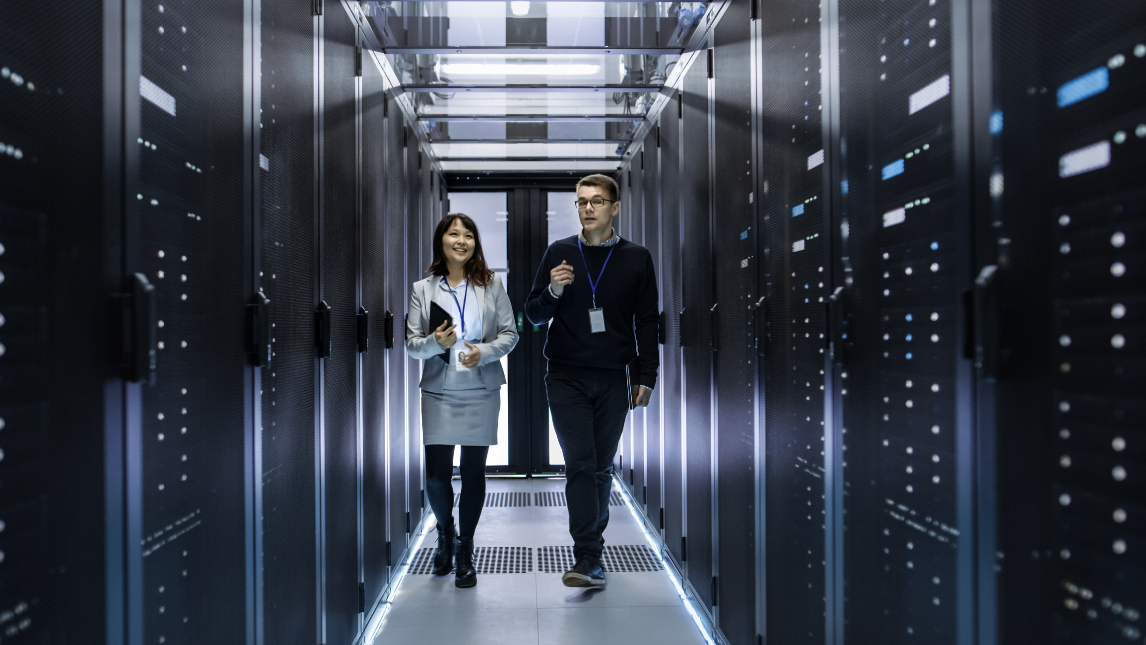 Caucasian Male And Asian Female It Technicians Walking Through Corridor Of Data Center With Rows Of Rack Servers. They Have Discussion, She Holds Tablet Computer.